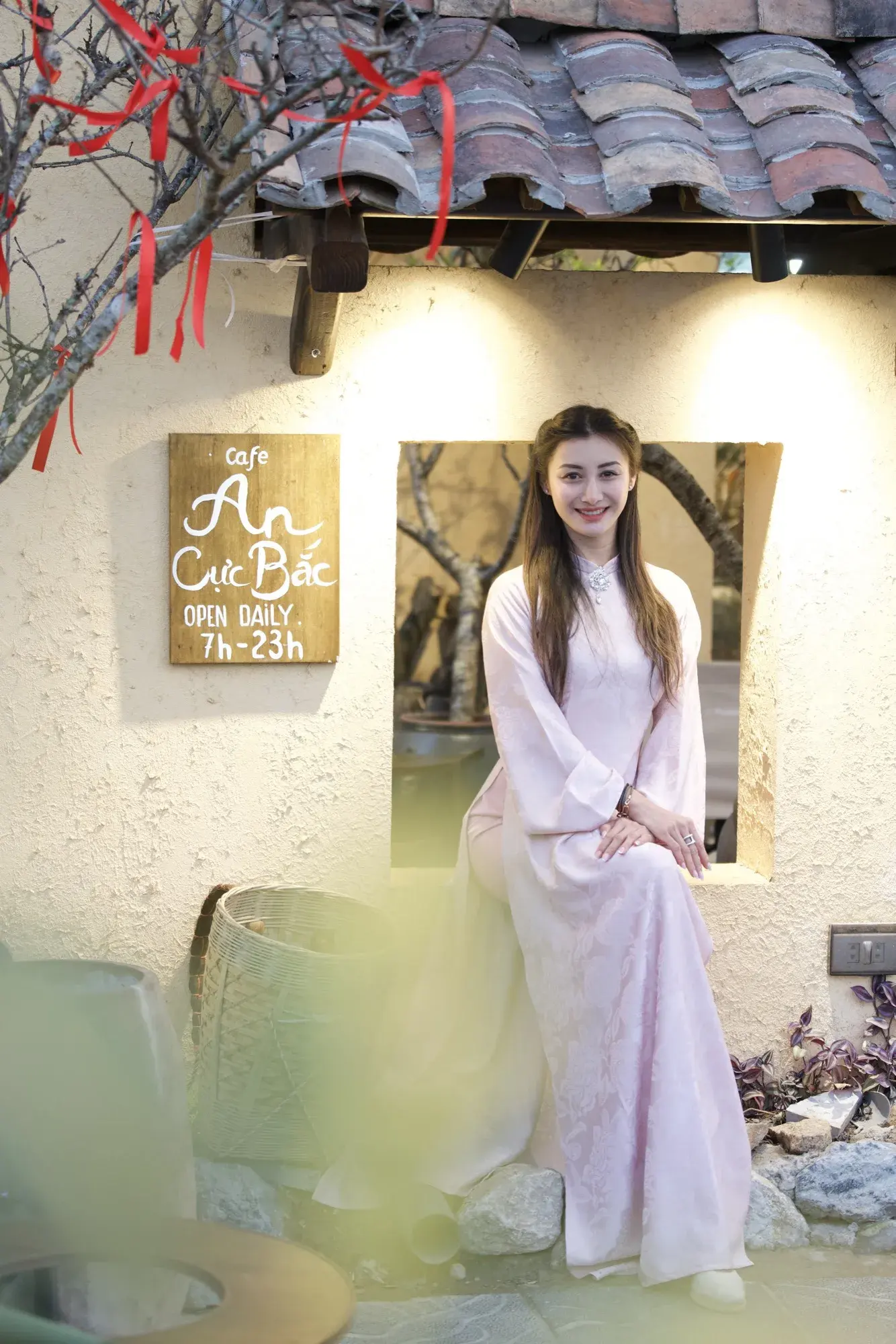 En robe traditionnelle rose pâle, souriante dans l’encadrement d’une fenêtre de café