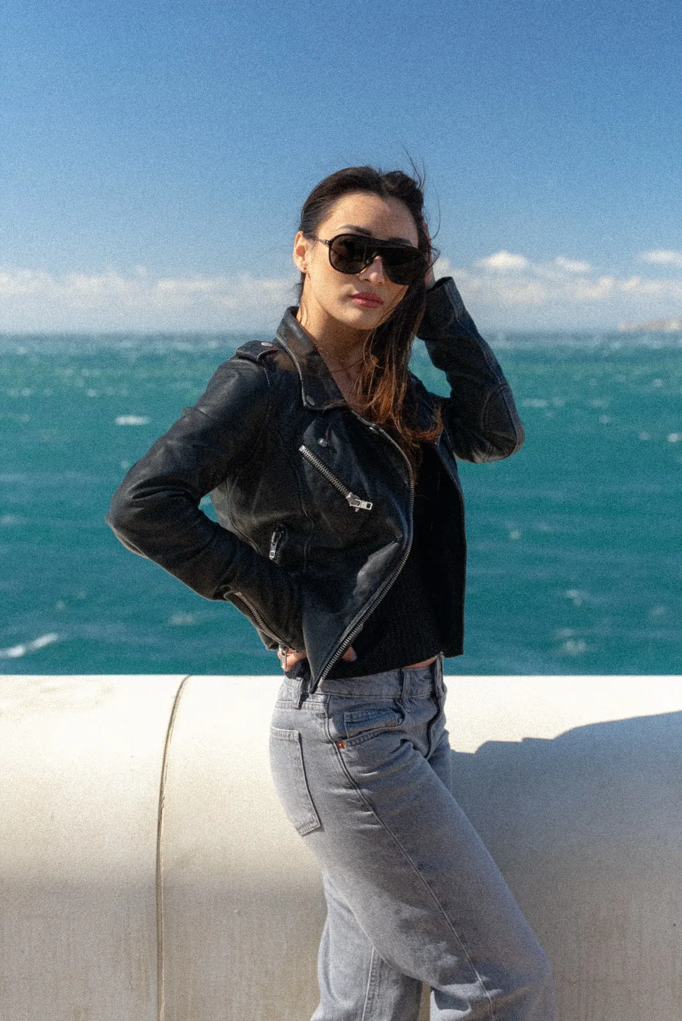 Appuyée contre un parapet blanc devant une mer turquoise, perfecto en cuir