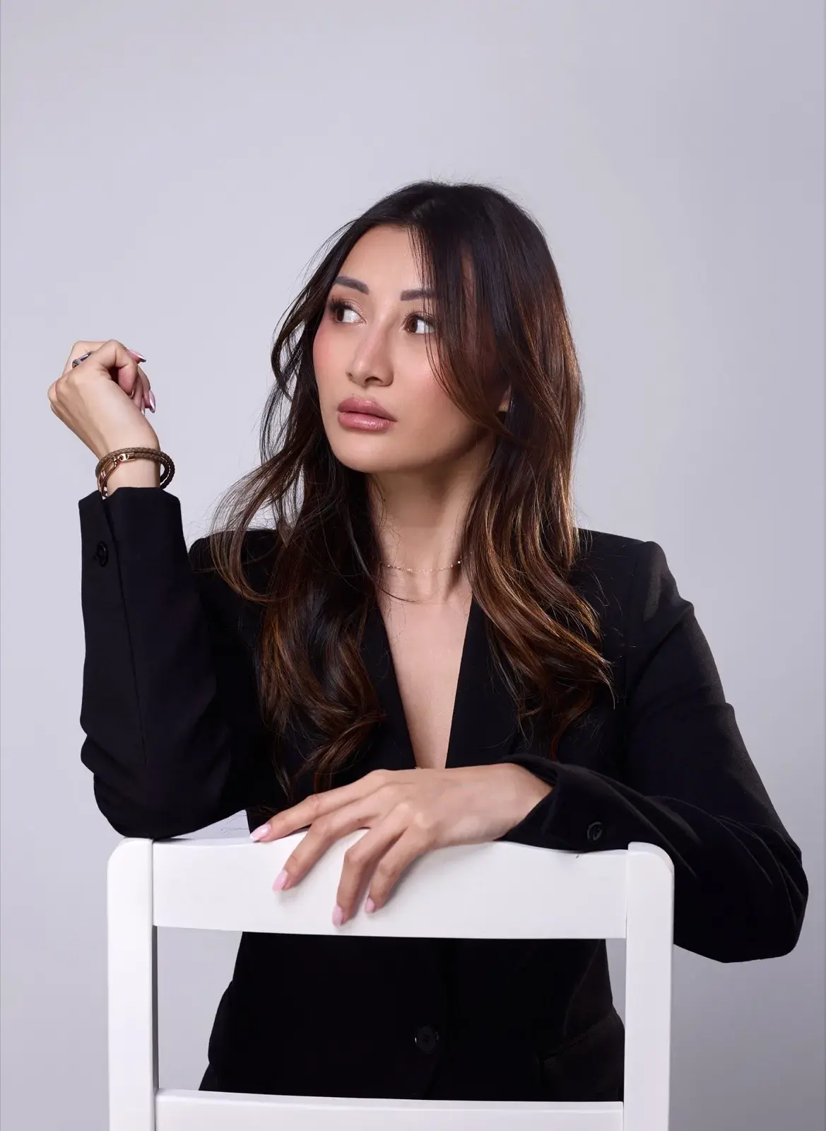 En blazer noir, accoudée au dossier d’une chaise, regard hors champ